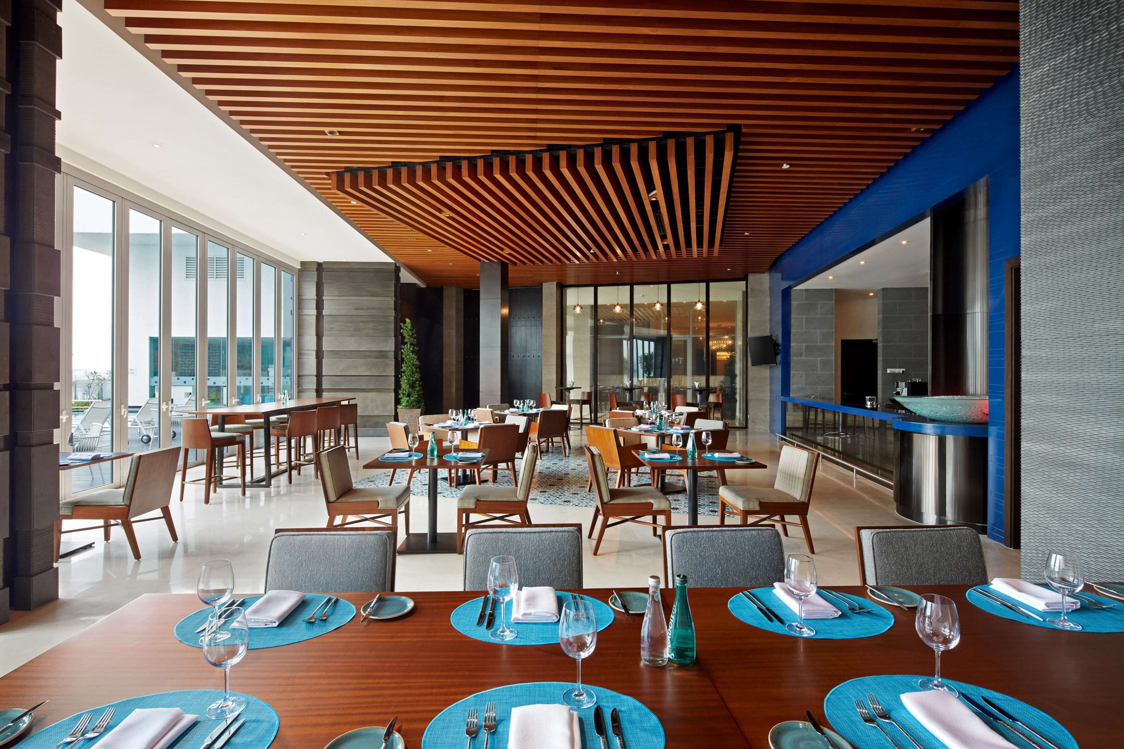 A modern dining area with wooden ceiling panels, blue placemats, and a view of the city through large windows.