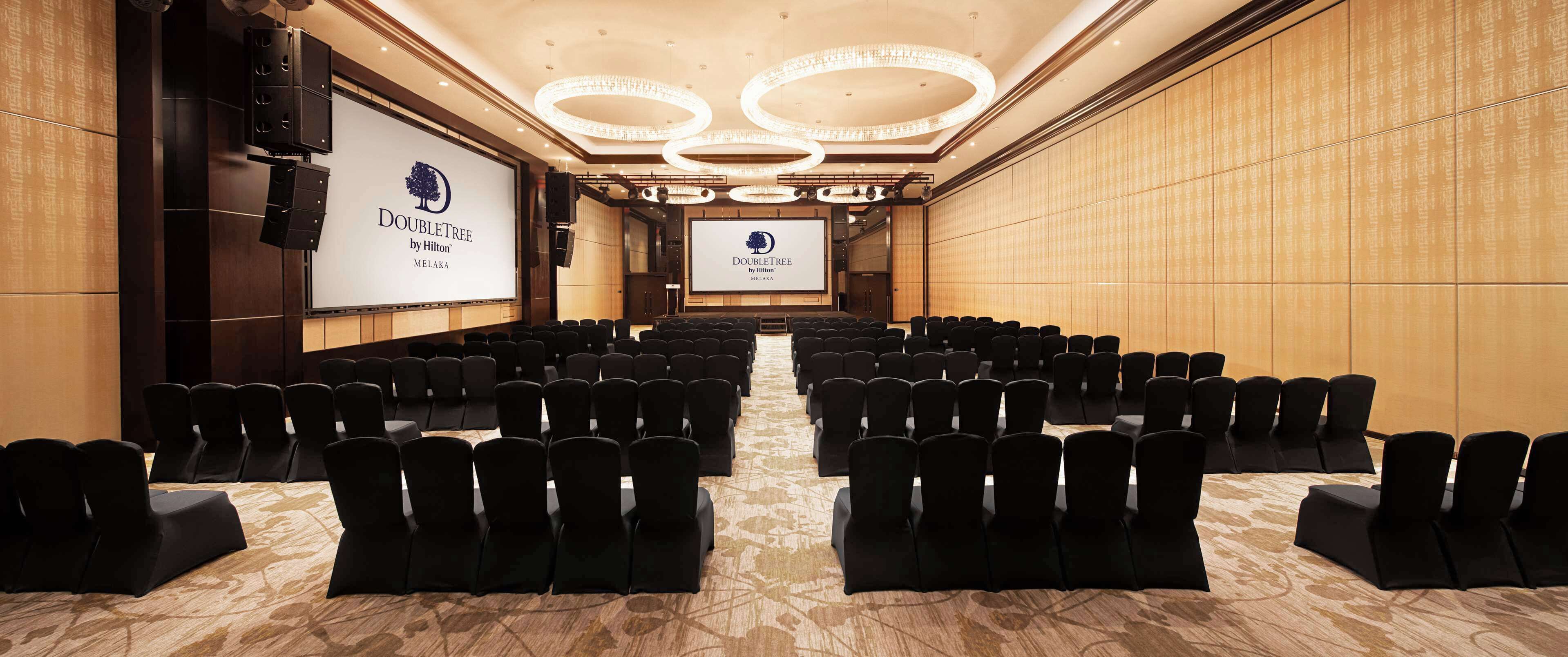 A conference room with rows of black chairs, a large screen displaying the DoubleTree by Hilton logo, and a chandelier on the ceiling.
