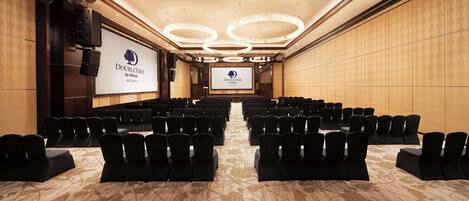 A conference room with rows of black chairs, a large screen displaying the DoubleTree by Hilton logo, and a chandelier on the ceiling.