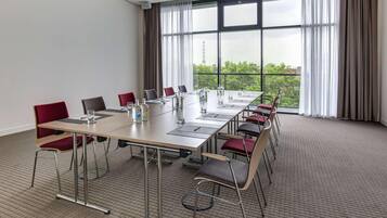 A conference room with a long table, red chairs, and large windows offering a view of greenery and buildings.