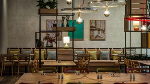 A restaurant with wooden tables and chairs, a high ceiling with hanging lights, and shelves with plants and decorative items. - IntercityHotel Duisburg (Duisburg)