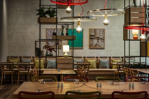 A restaurant with wooden tables and chairs, a high ceiling with hanging lights, and shelves with plants and decorative items. - IntercityHotel Duisburg (Duisburg)