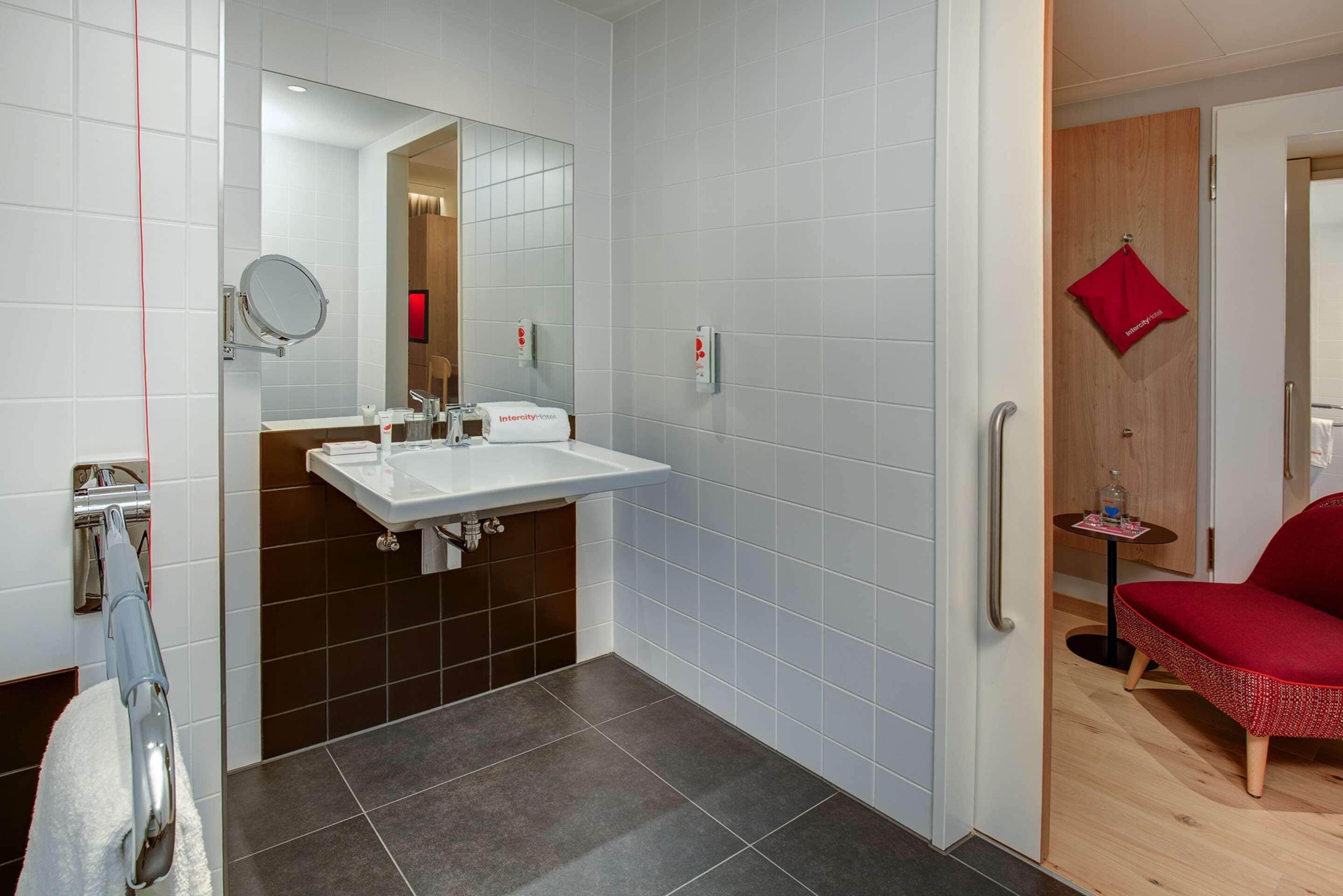 Een moderne badkamer met een witte wastafel, donkere tegels, een spiegel en een rode handdoek aan een deur.