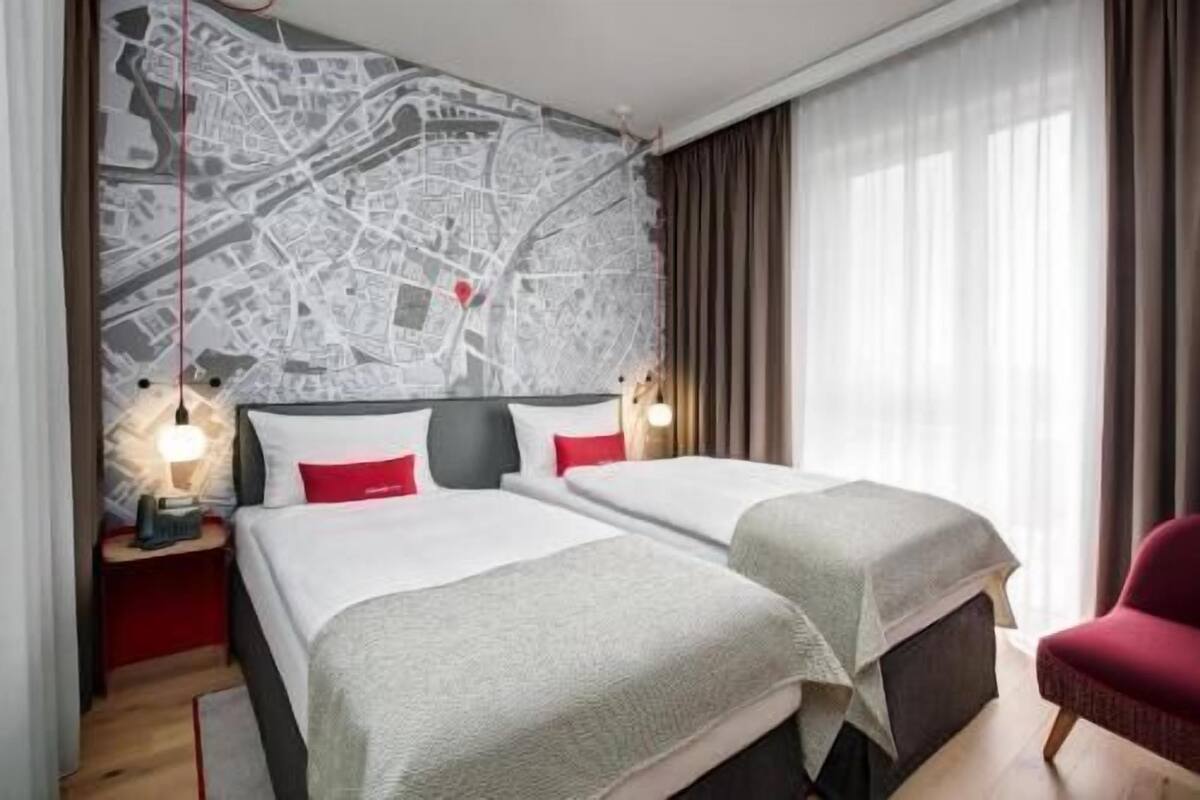 A hotel room with two beds, a map on the wall, a red chair, and a nightstand with a lamp.