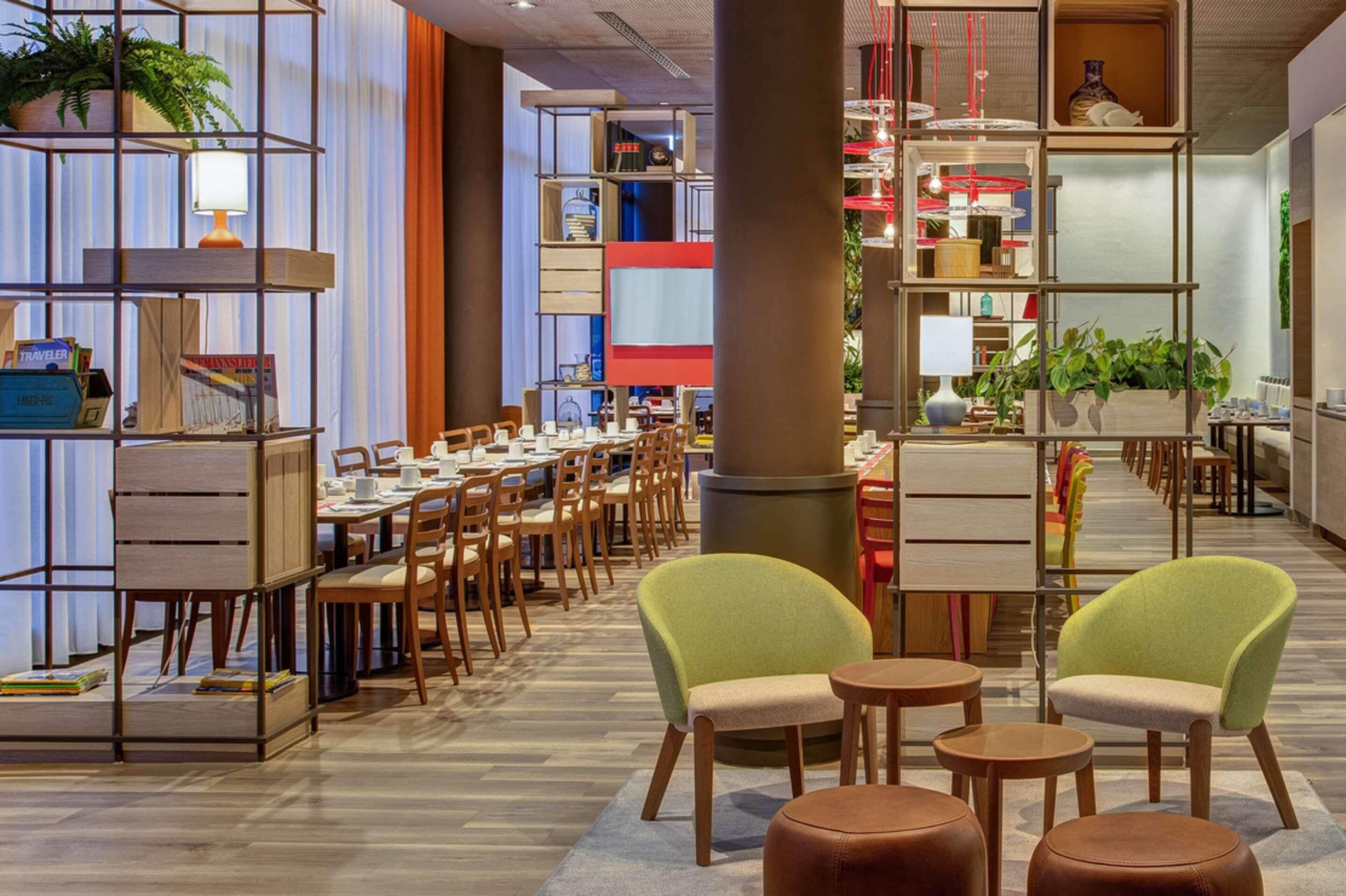 A modern restaurant with multiple dining areas, wooden furniture, and shelves with books and plants.