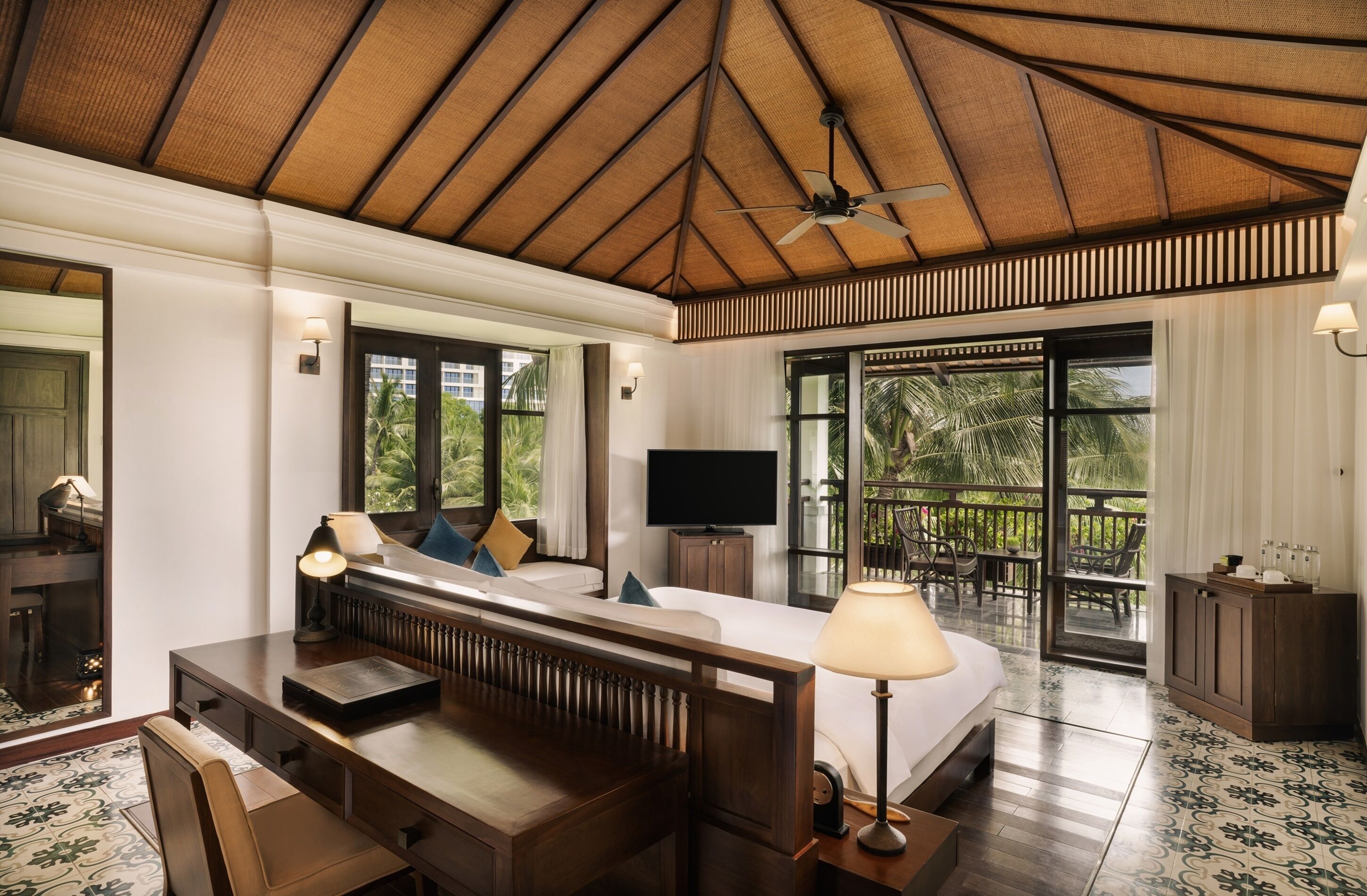 A hotel room with a large bed, a desk, a television, and a balcony with outdoor furniture.