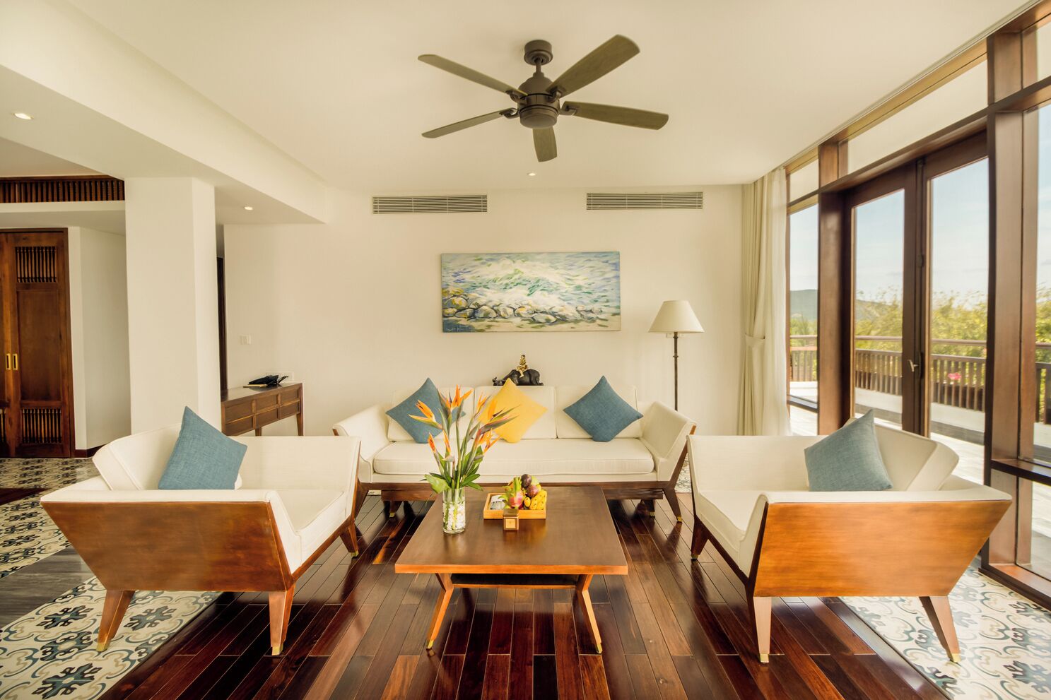 A spacious living room with a ceiling fan, a painting, and a view of the outdoors through large windows.