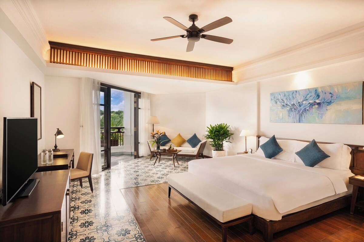 A hotel room with a large bed, a desk with a TV, a ceiling fan, and a painting on the wall.