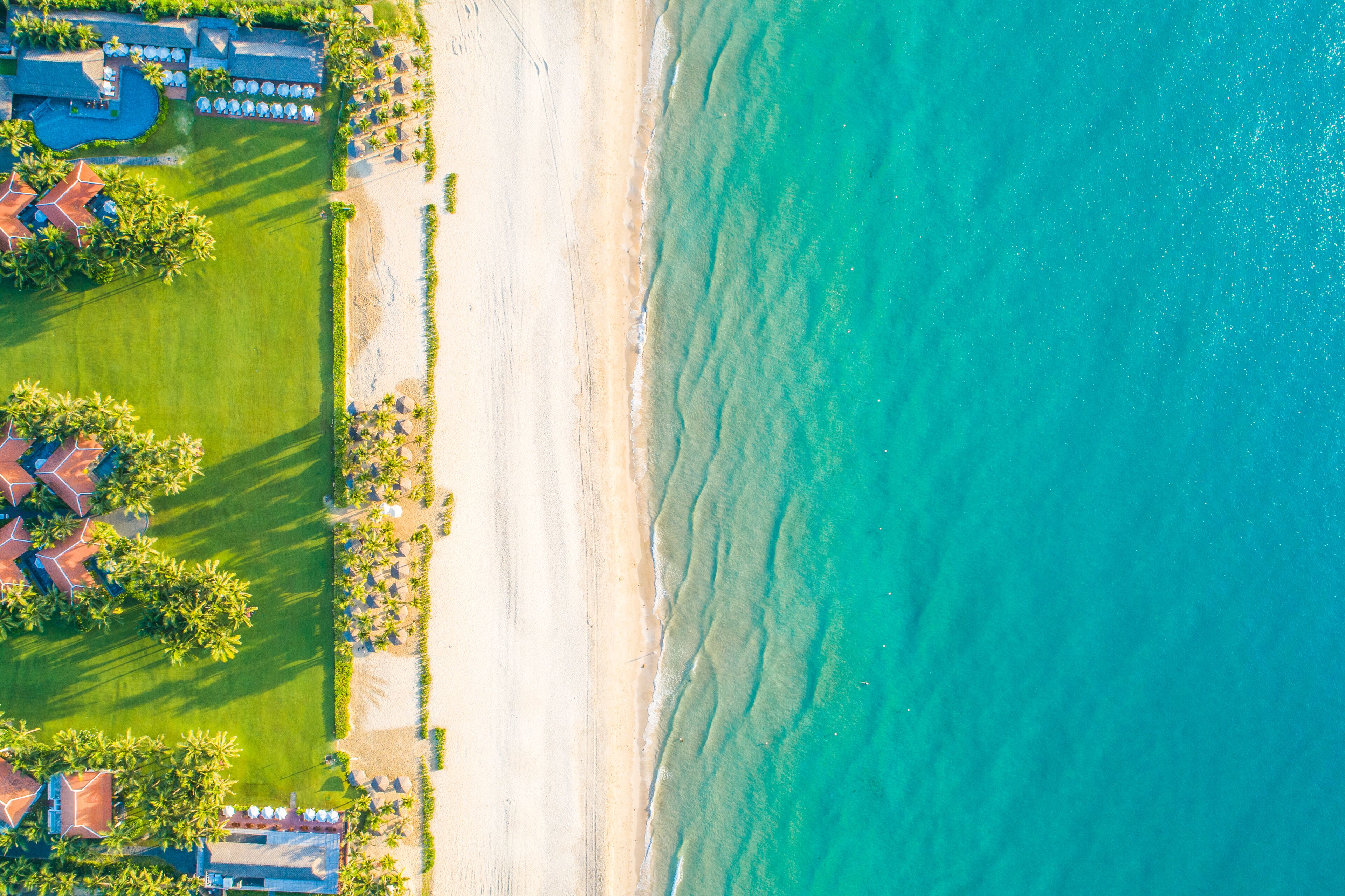 An aerial view of a beach resort with a sandy shoreline, clear turquoise waters, and a well-maintained lawn area with palm trees.
