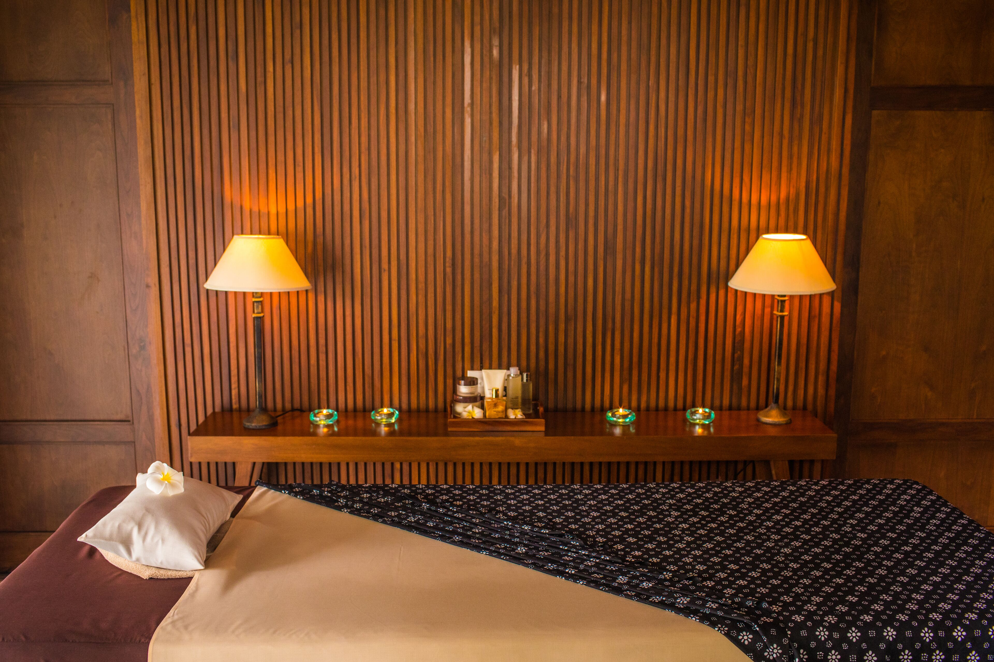 Ein Hotelzimmer mit Holzwand, einem Nachttisch aus Holz mit zwei Lampen, einem kleinen Tisch mit Toilettenartikeln und einem Bett mit gemustertem Überwurf.