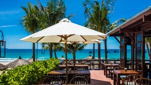 An outdoor dining area with wooden tables and chairs, surrounded by palm trees and a view of the ocean. - The Anam Cam Ranh (Cam Lam)