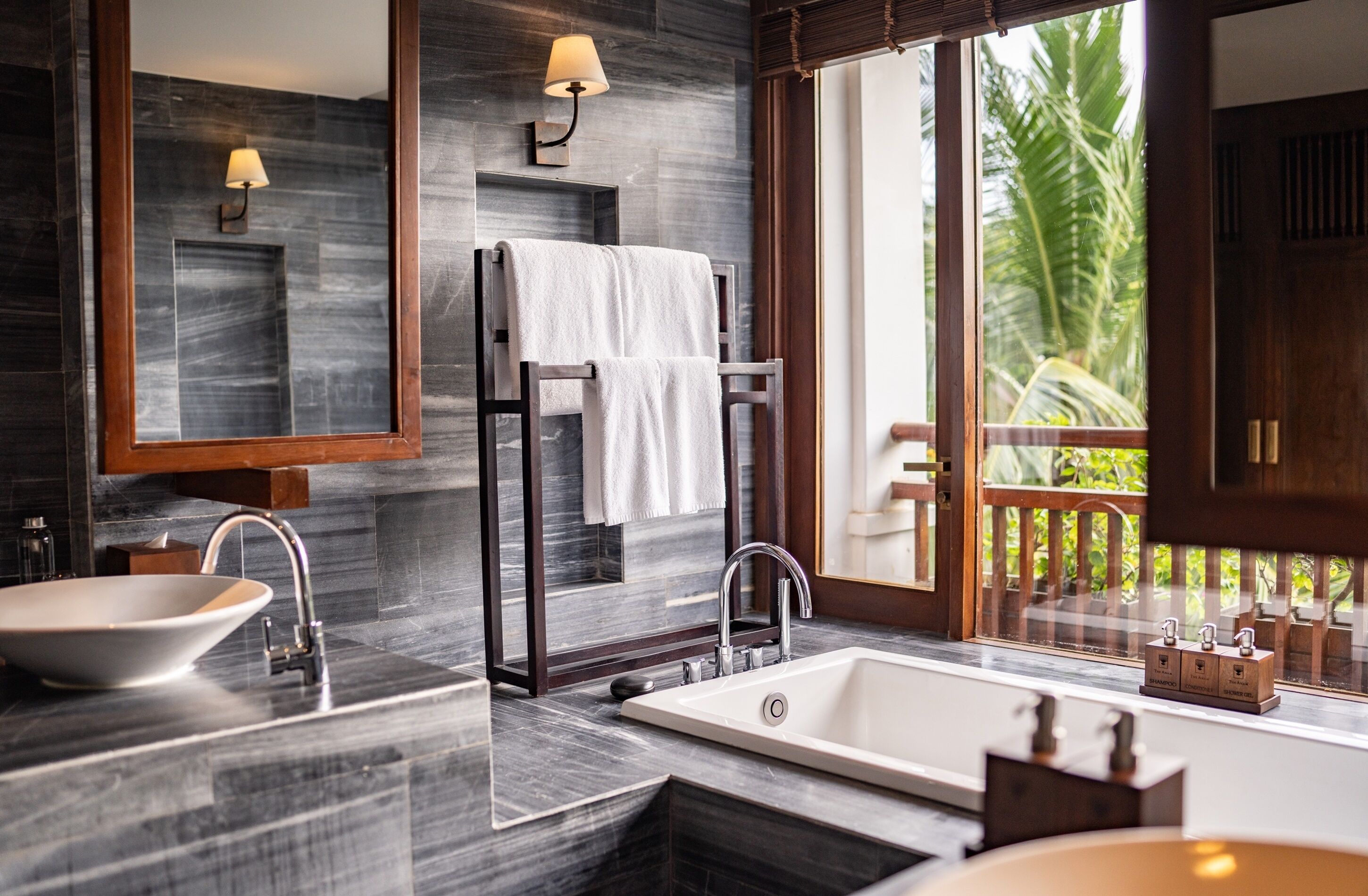 Un baño moderno con una bañera grande, un lavabo y un espejo. Hay un perchero con una toalla colgada, y una ventana con vista a palmeras.