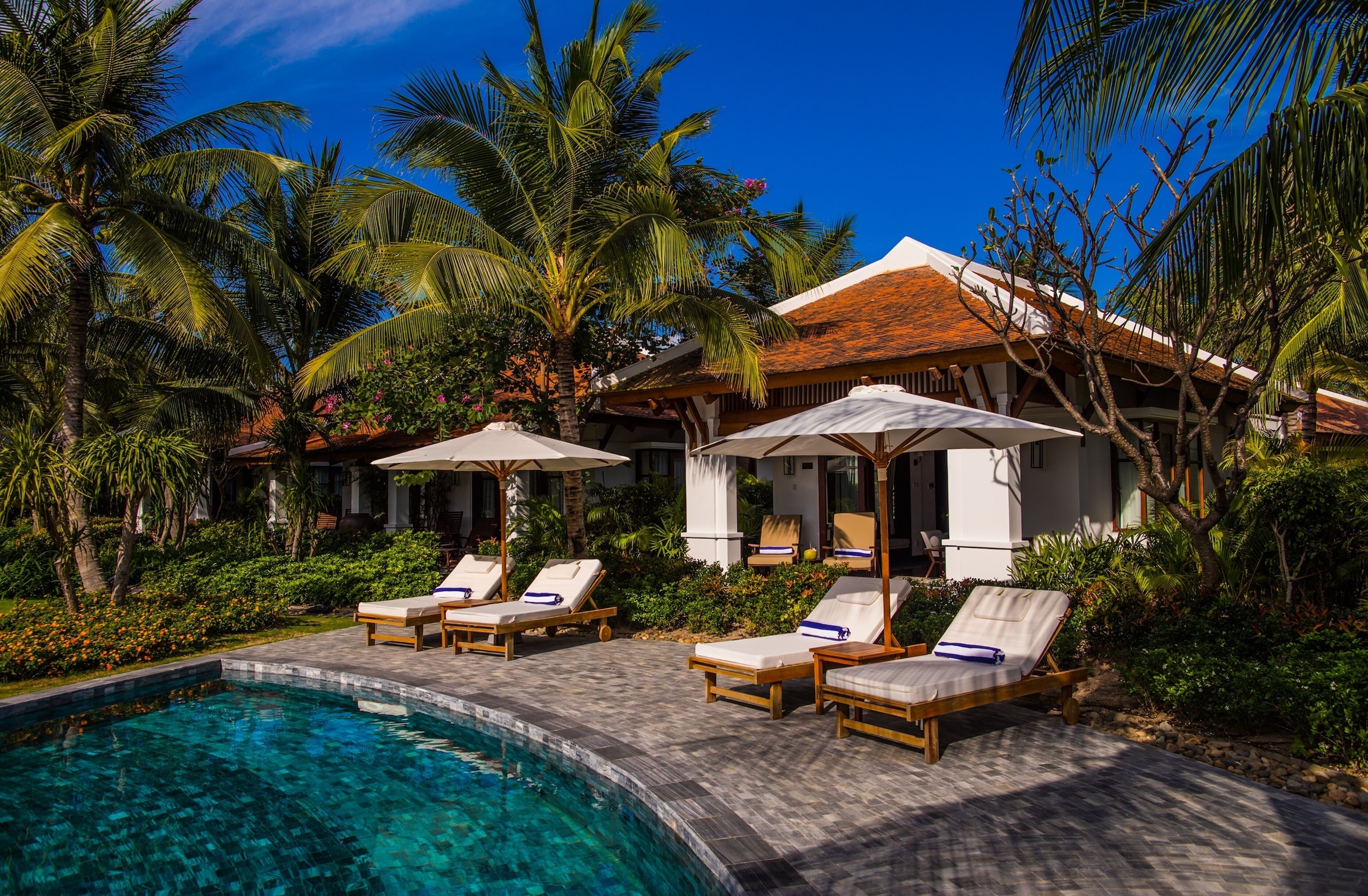 Zona junto a la piscina con sillones de descanso y sombrillas, rodeada de exuberante vegetación y palmeras.