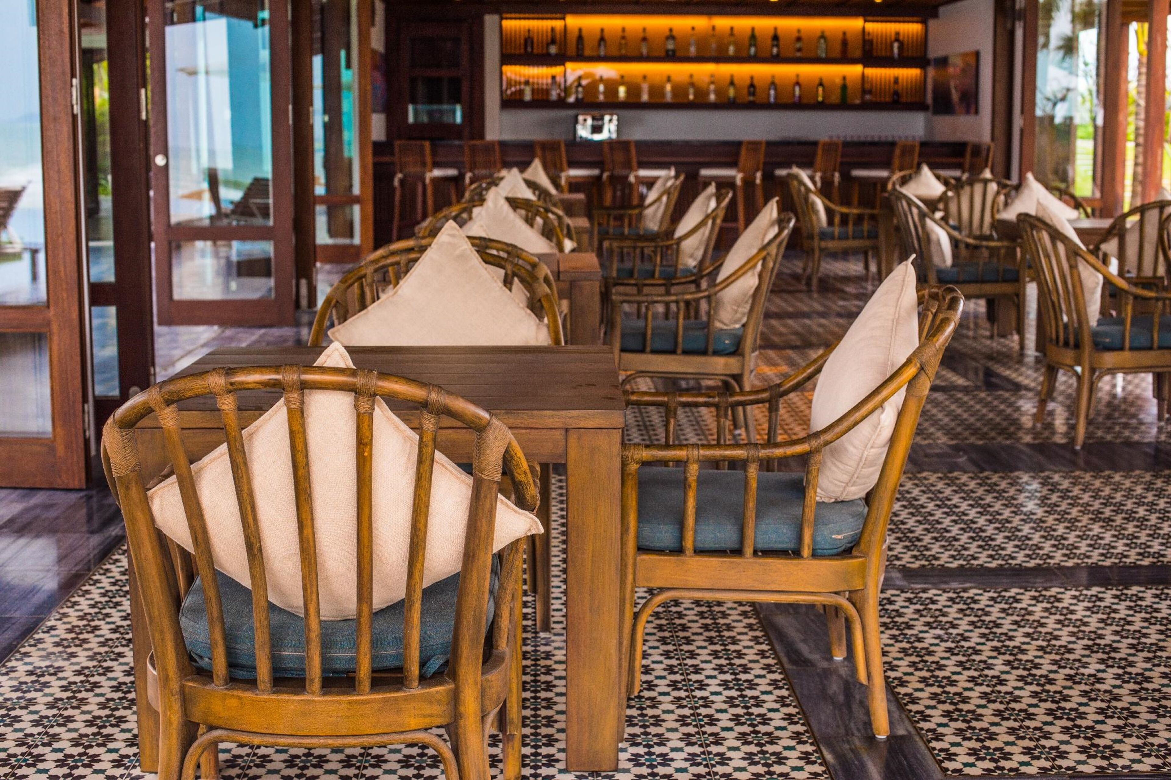 L’intérieur d’un restaurant est aménagé avec des chaises et des tables en bois, un sol à motifs et un bar où sont exposées des bouteilles.