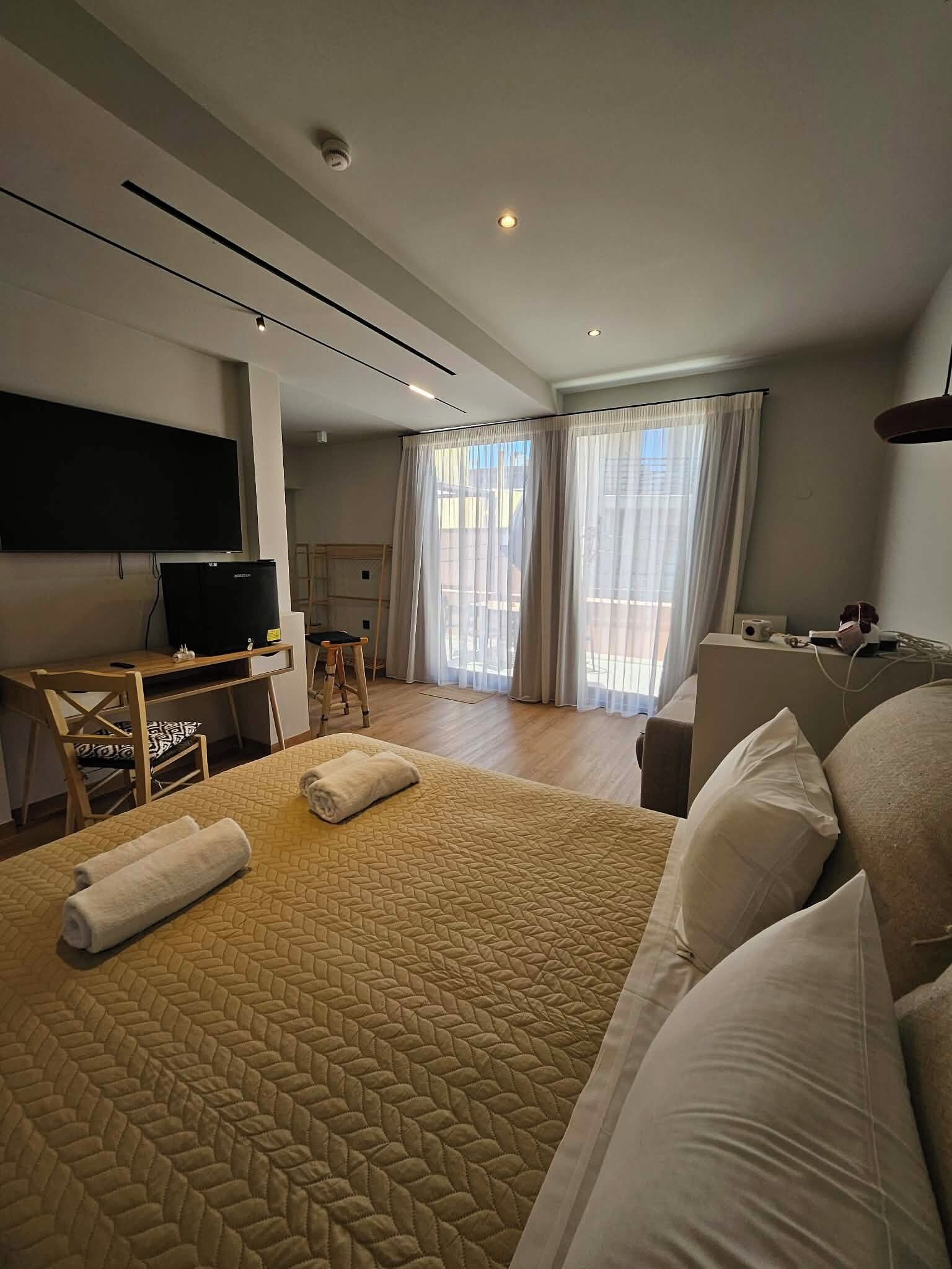Una habitación de hotel moderna con una cama grande, un escritorio con ordenador, un televisor de pantalla plana y un balcón con cortinas.