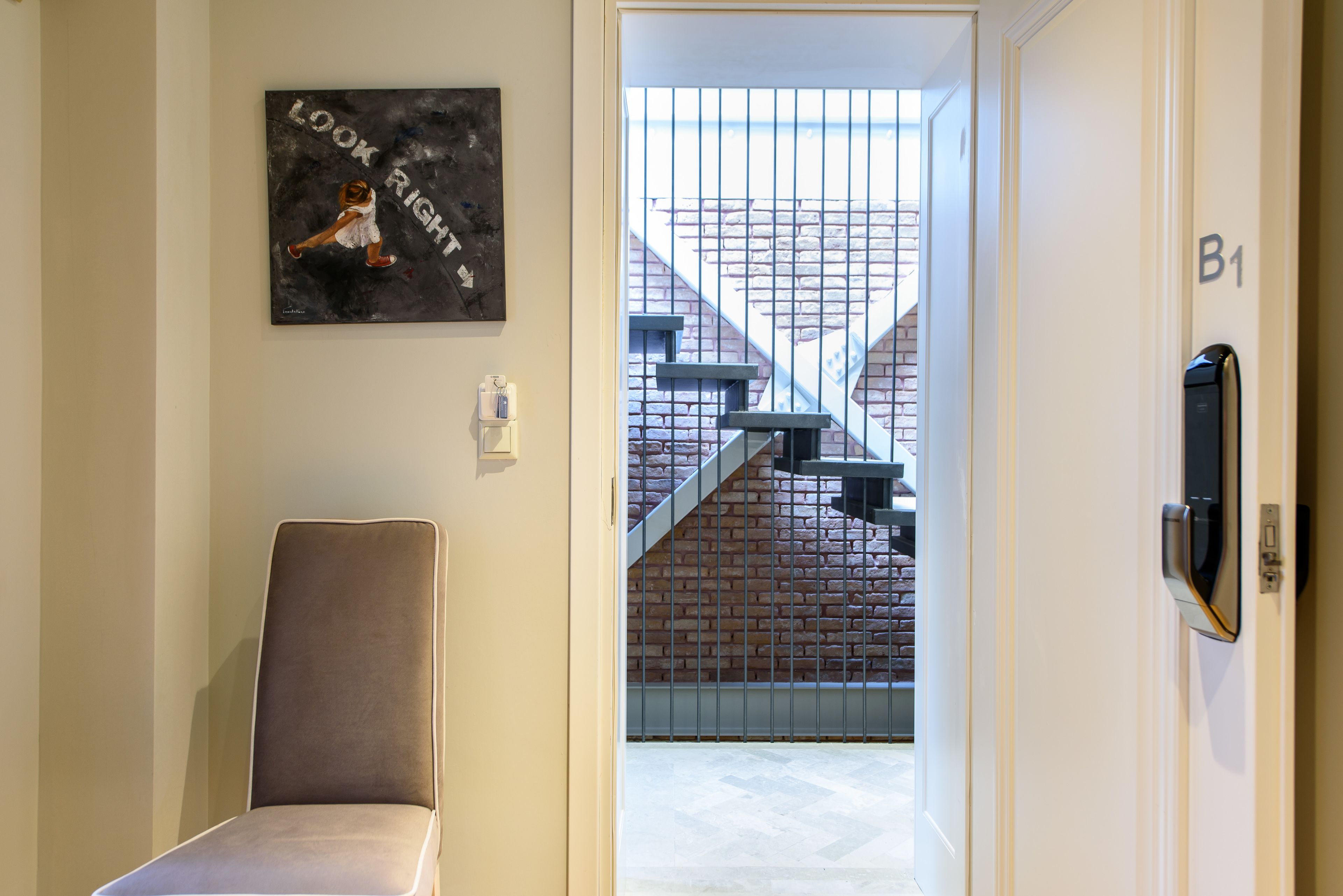 A hallway with a chair, a painting, a door labeled B1, and a staircase leading to a brick wall.