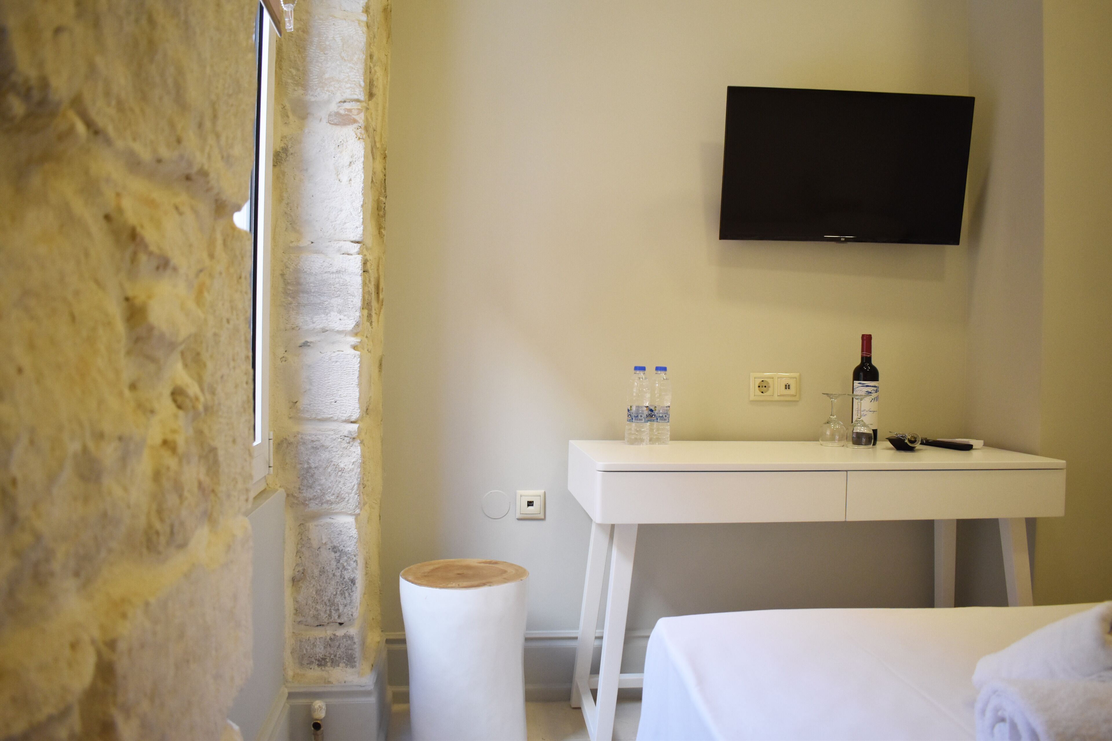 A hotel room with a white bed, a white desk with a flat-screen TV, a bottle of wine, and two water bottles.