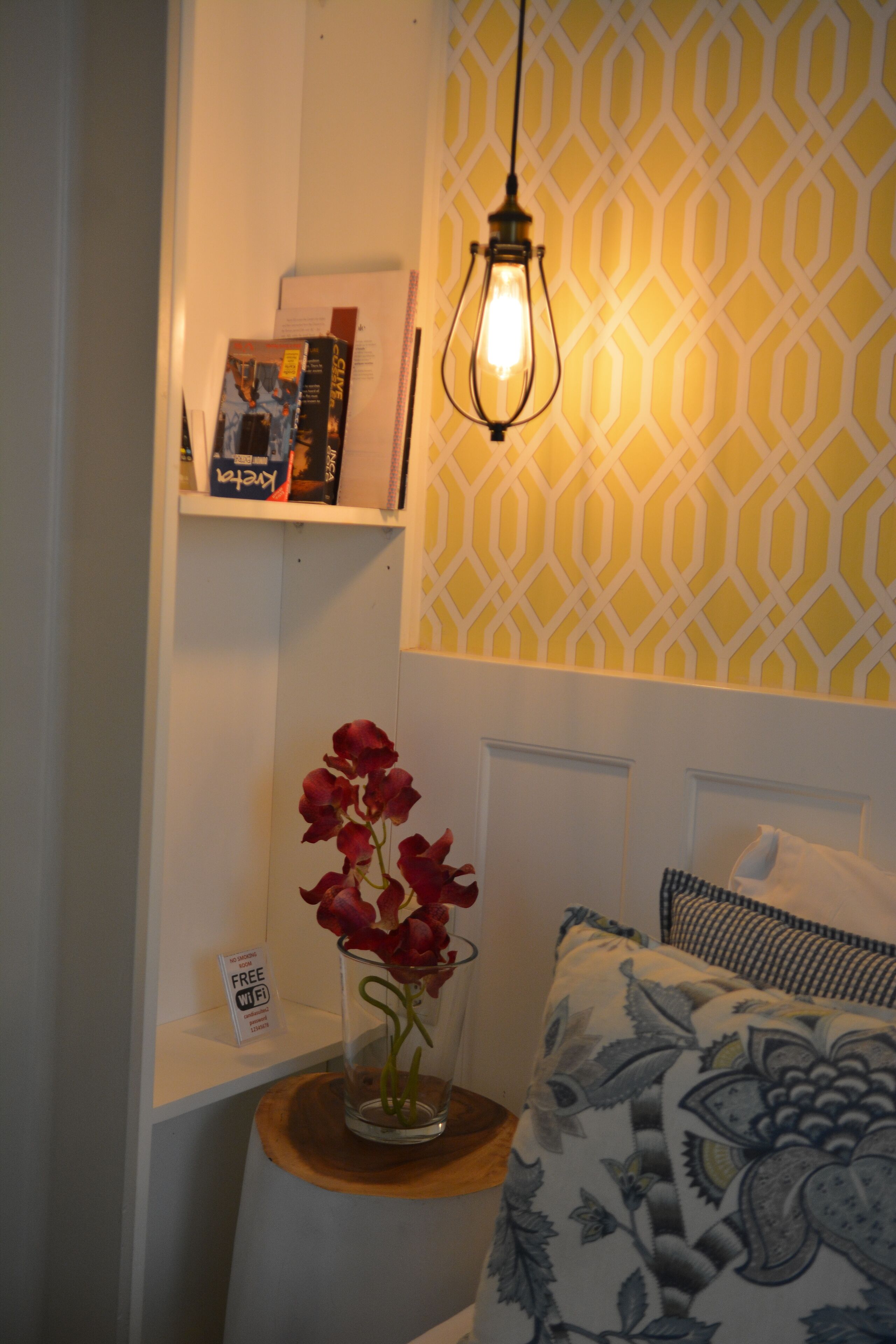 A room with a bed, a bedside table with a vase of flowers, a shelf with books and a sign, and a patterned wall.