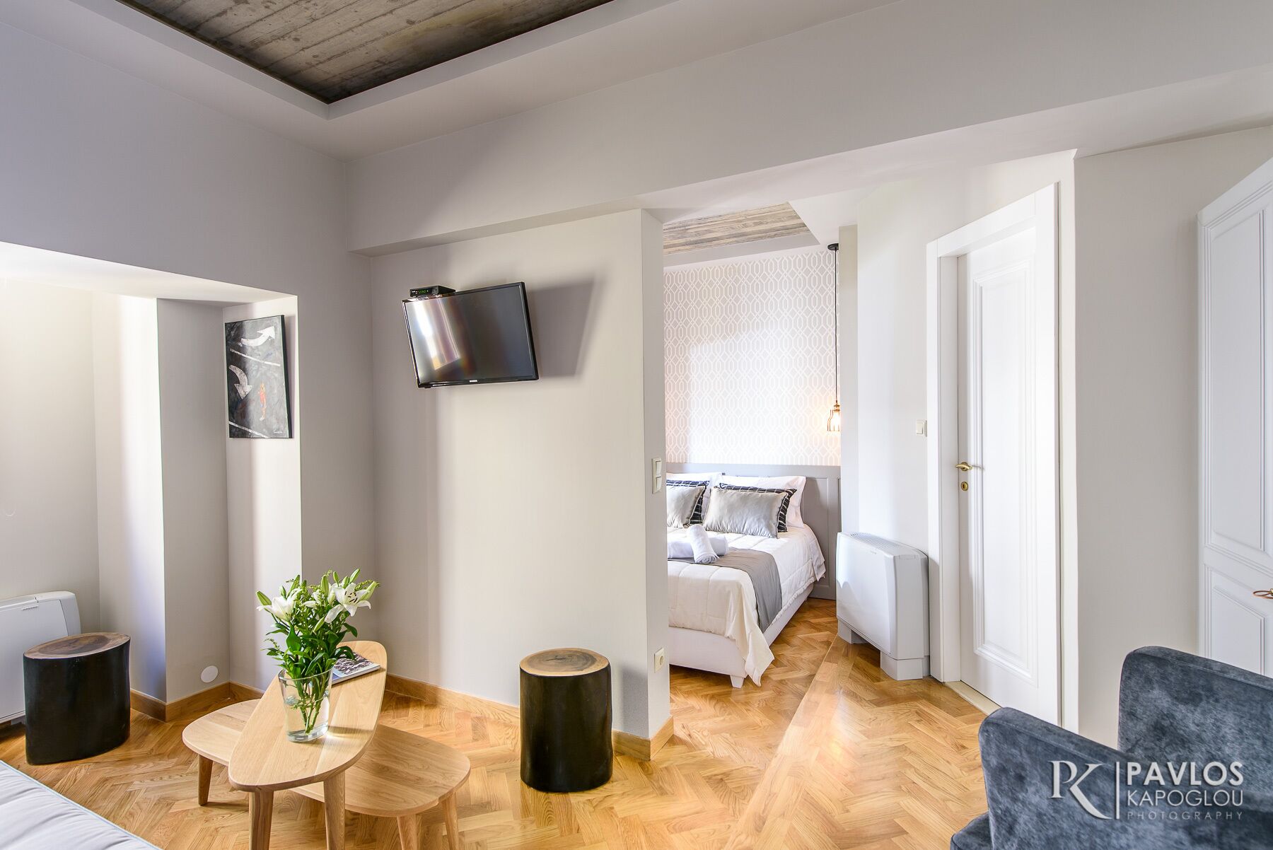 Una habitación de hotel moderna con piso de madera, una cama con ropa de cama blanca, una mesita con sillas y un televisor montado en la pared.