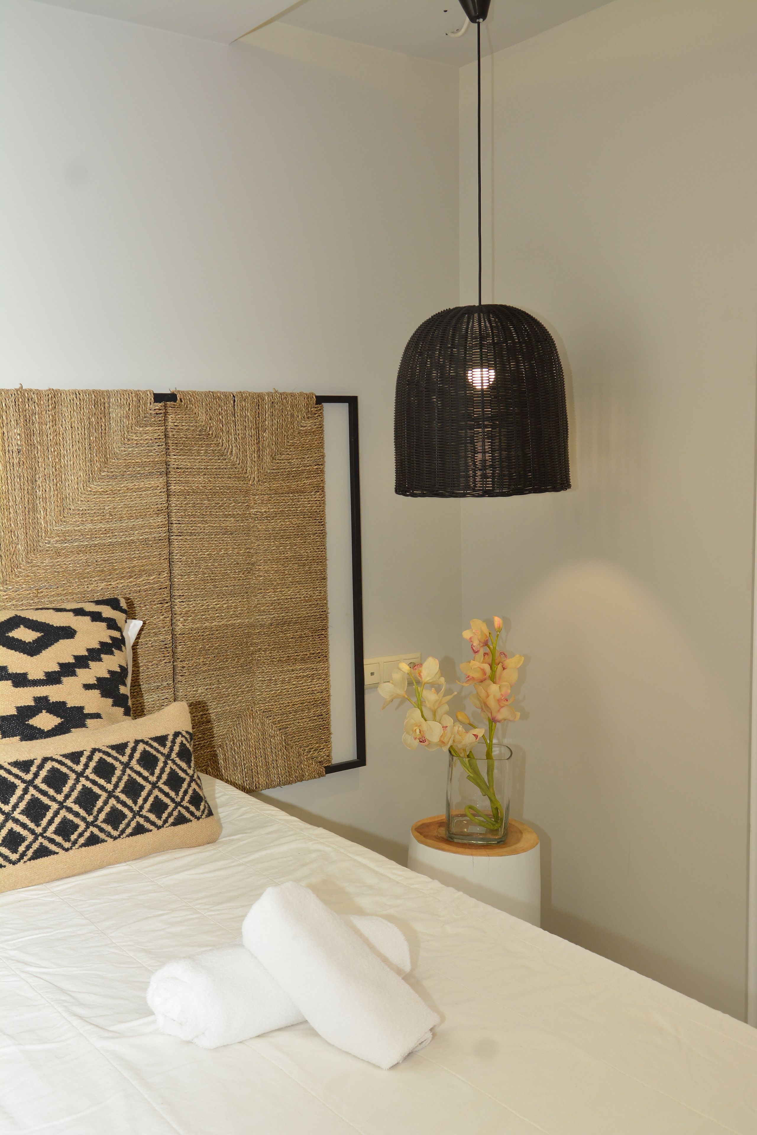 A neatly made bed with white linens and two rolled towels, a woven headboard, a black pendant light, and a vase with flowers on a bedside table.