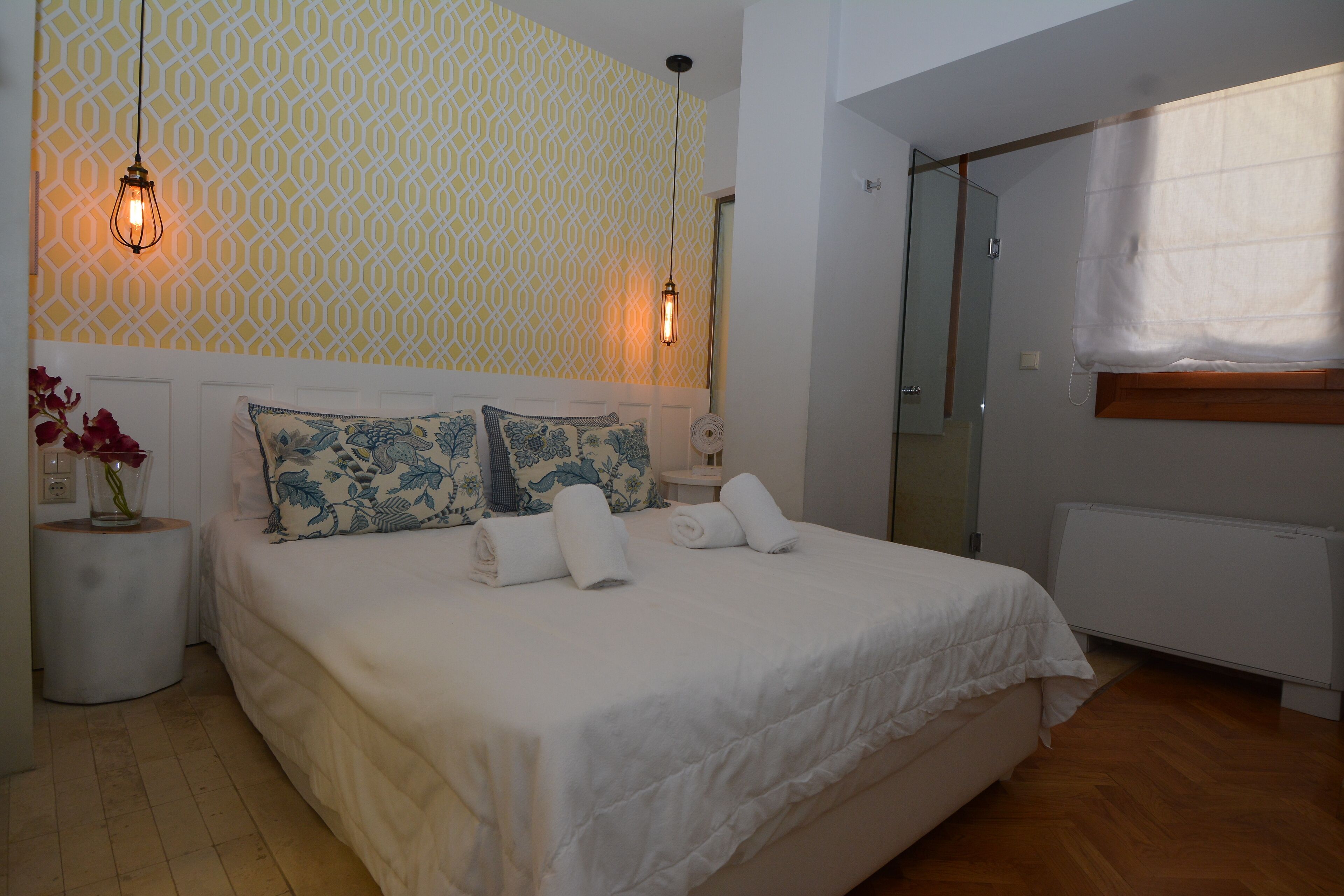 A neatly made bed with white linens and blue floral pillows, a bedside table with a vase of flowers, and a patterned wall.