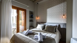 Un dormitorio con cama, almohadas, mesita de noche y ventana con cortinas.