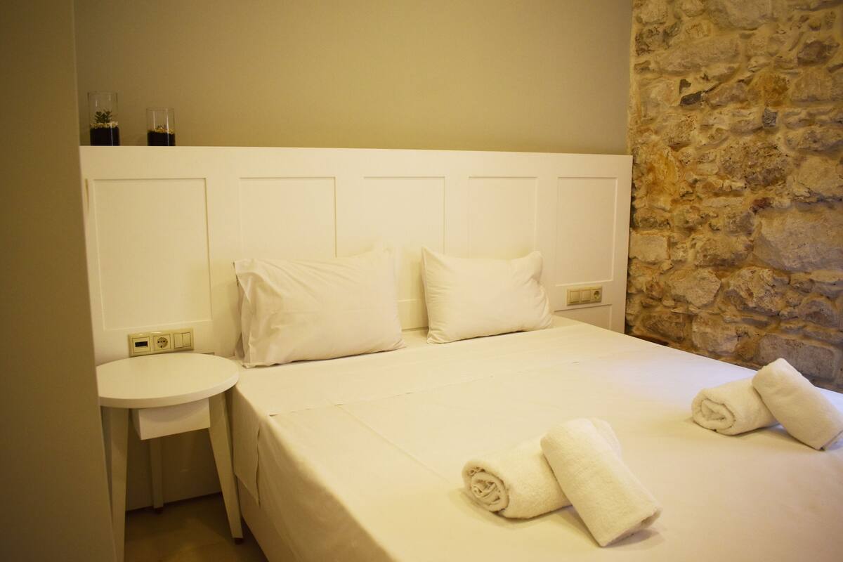 Une chambre d’hôtel avec un lit, deux oreillers, une table de chevet ronde et un mur en pierre.