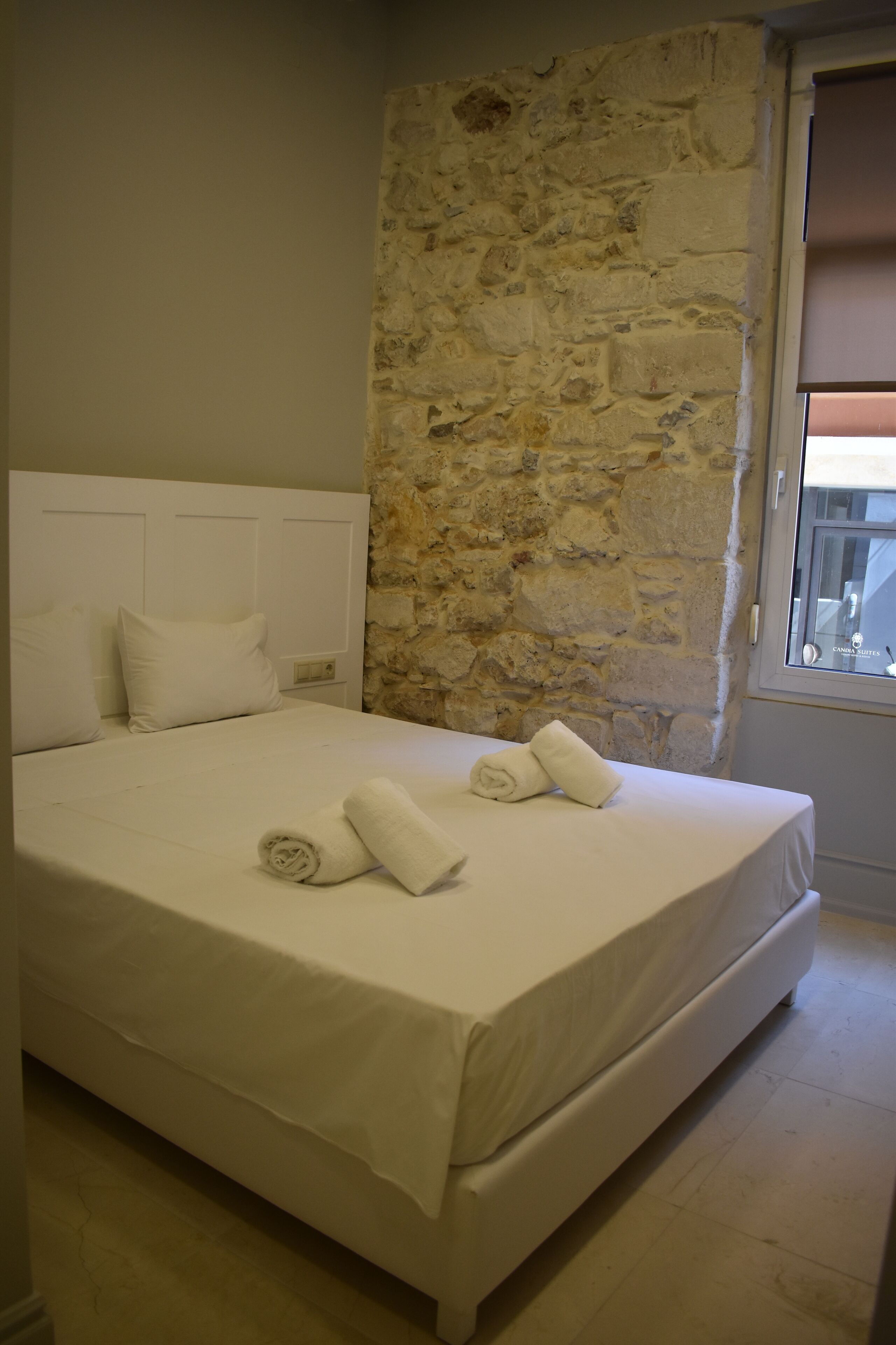 A neatly made bed with white linens and two rolled towels, a stone wall, and a window with blinds.