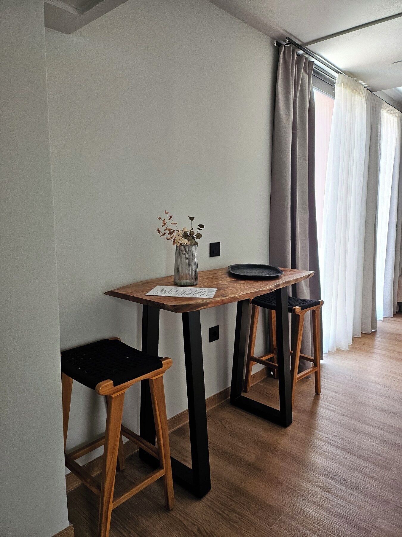 Un petit coin repas avec une table en bois, deux tabourets noirs et un vase contenant des fleurs séchées.