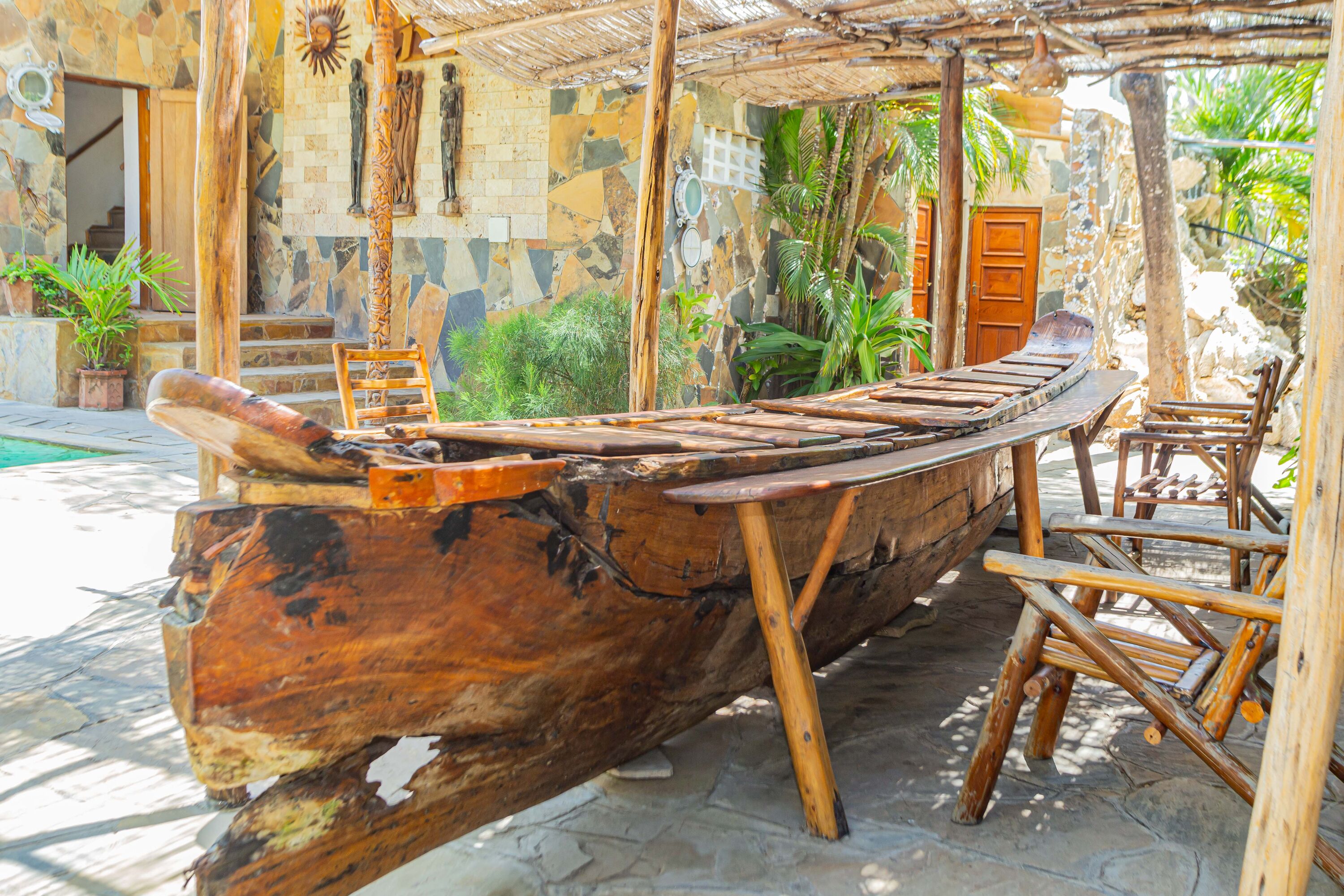 A wooden boat-shaped bench with a rustic design, surrounded by stone steps and wooden chairs, set in a garden with potted plants and a stone wall.