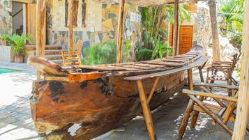 A wooden boat-shaped bench with a rustic design, surrounded by stone steps and wooden chairs, set in a garden with potted plants and a stone wall.