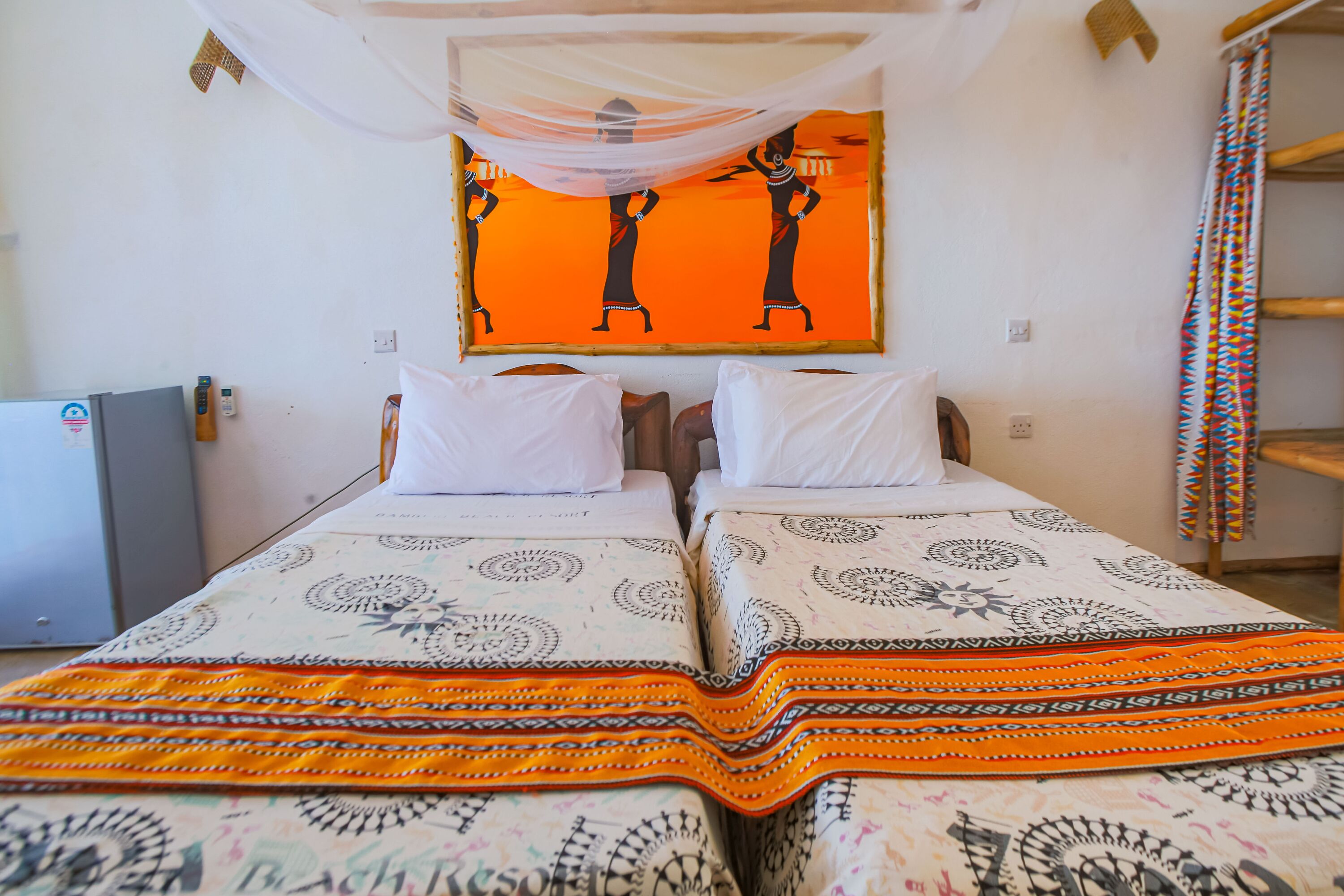 A room with two beds, each with patterned bedding and white pillows. There is a wall-mounted artwork above the beds and a small desk with a chair to the left.