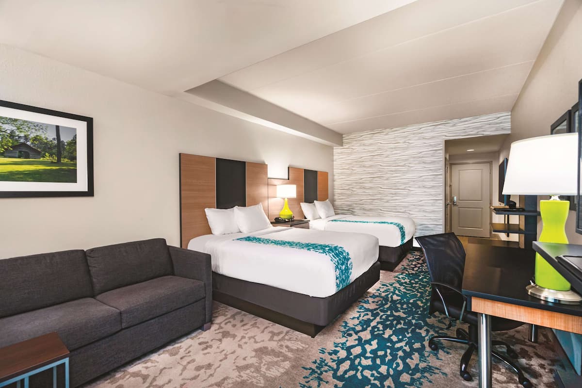 A hotel room with two beds, a desk, a chair, a couch, and a framed picture on the wall.