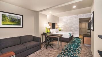 A hotel room with a gray sofa, a desk with two lamps, a bed with a white headboard, and a patterned carpet.