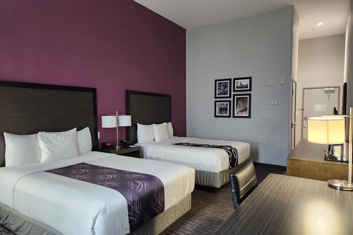 A hotel room with two beds, a desk, a chair, a nightstand, a lamp, and framed pictures on the wall.