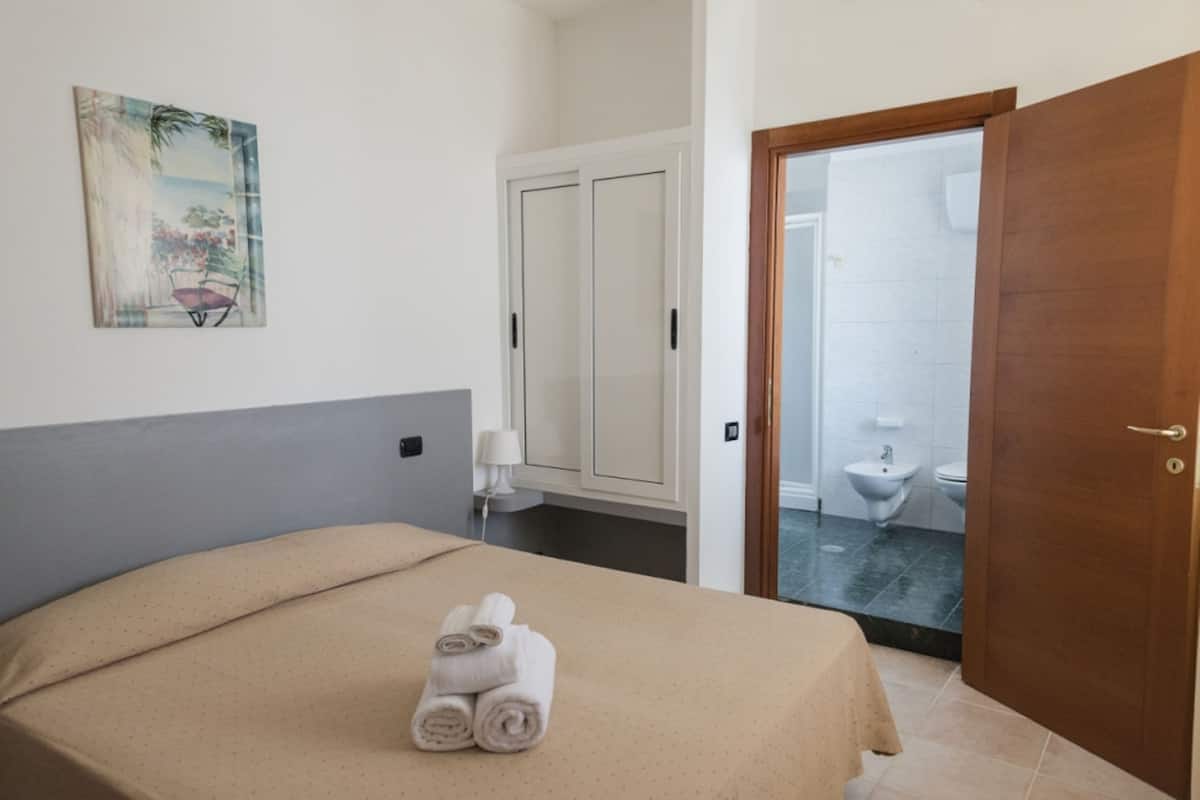 Una habitación de hotel con cama, un cuadro, un armario y un baño visible a través de una puerta abierta.