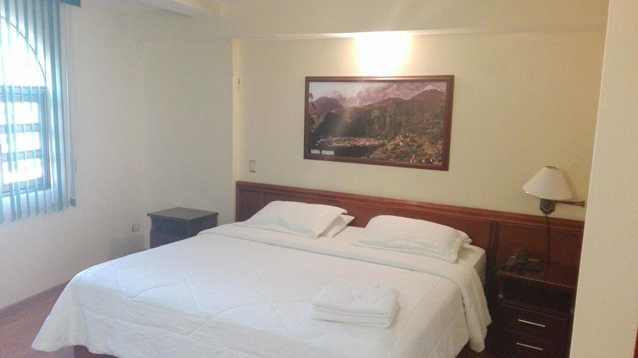 Un lit bien fait, avec du linge de lit blanc, une table de chevet avec une lampe et, au mur, un tableau représentant un paysage montagneux.