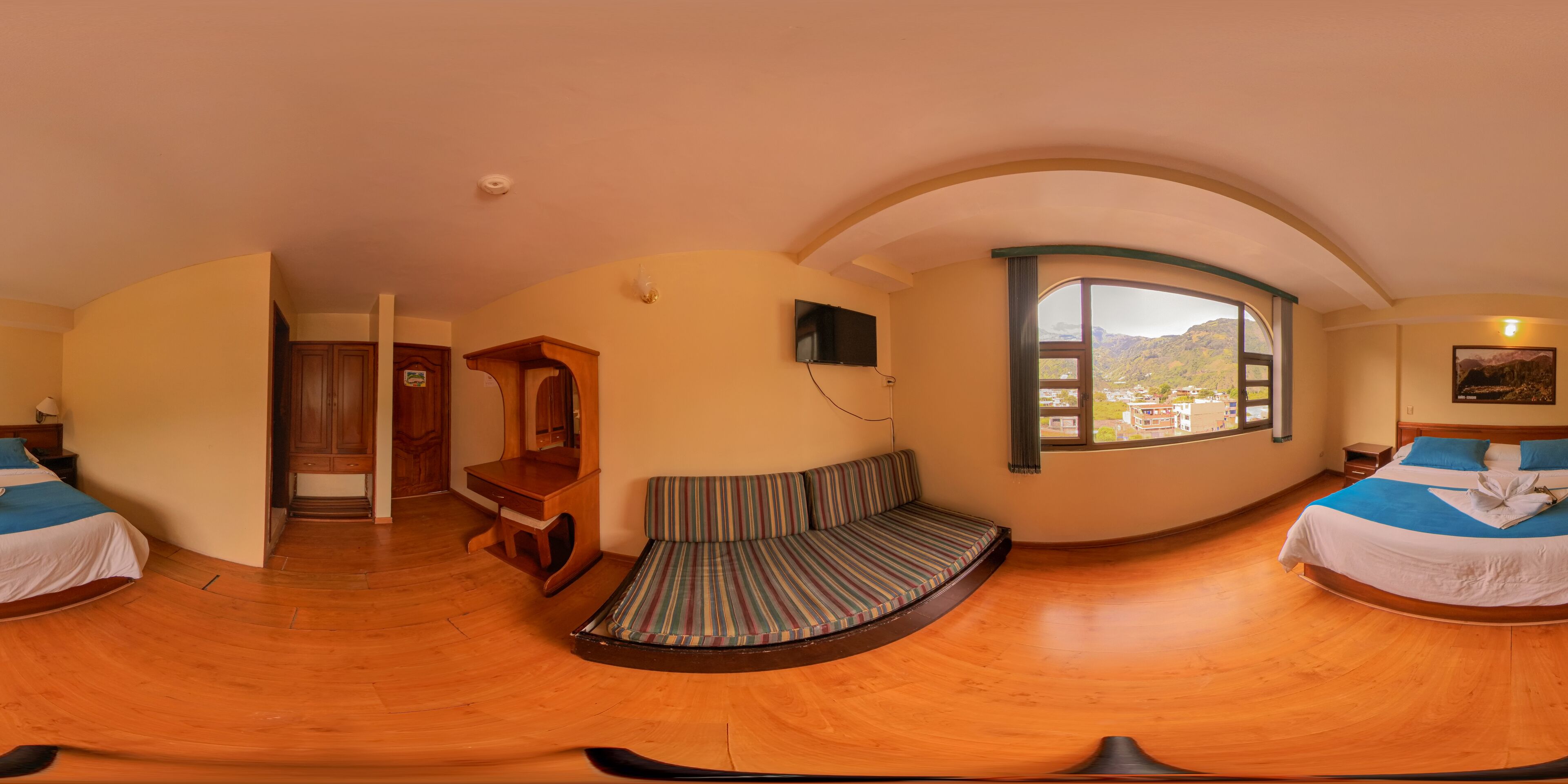 A room with a curved ceiling, a bed with a striped mattress, a wooden desk, a television, and a window with a view of mountains.