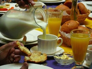 Une table mise pour le petit-déjeuner, avec une carafe d’où l’on verse du lait, un verre de jus d’orange et un panier de pain.