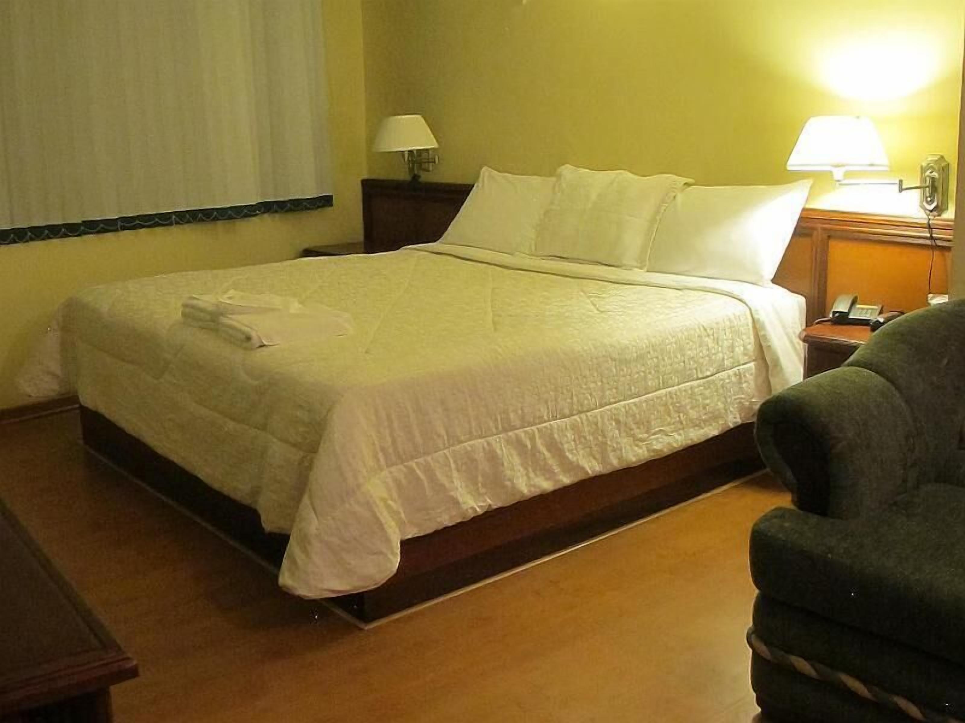 A hotel room with a bed, a chair, a nightstand with a lamp, and a wall-mounted phone.