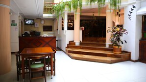 Un vestíbulo de hotel con mostrador de recepción, zona de comedor con mobiliario de madera, una escalera que sube a un nivel superior y plantas en macetas.
