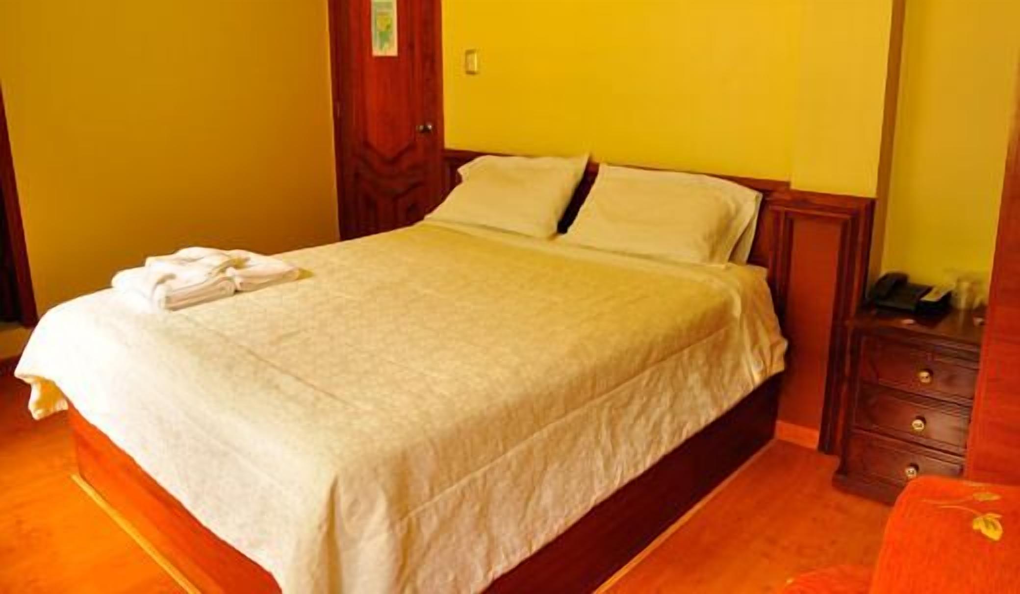 Una cama bien hecha con edredón blanco y almohadas, una mesita de noche de madera con un teléfono y un objeto pequeño encima, y una puerta de madera con un cartel.