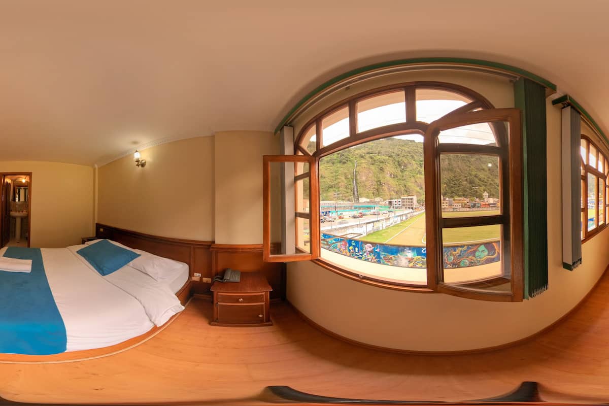 Een kamer met een groot rondboogvenster, een bed met een blauw-witte spreidekker, een klein houten nachtkastje en uitzicht op een groen landschap en gebouwen.