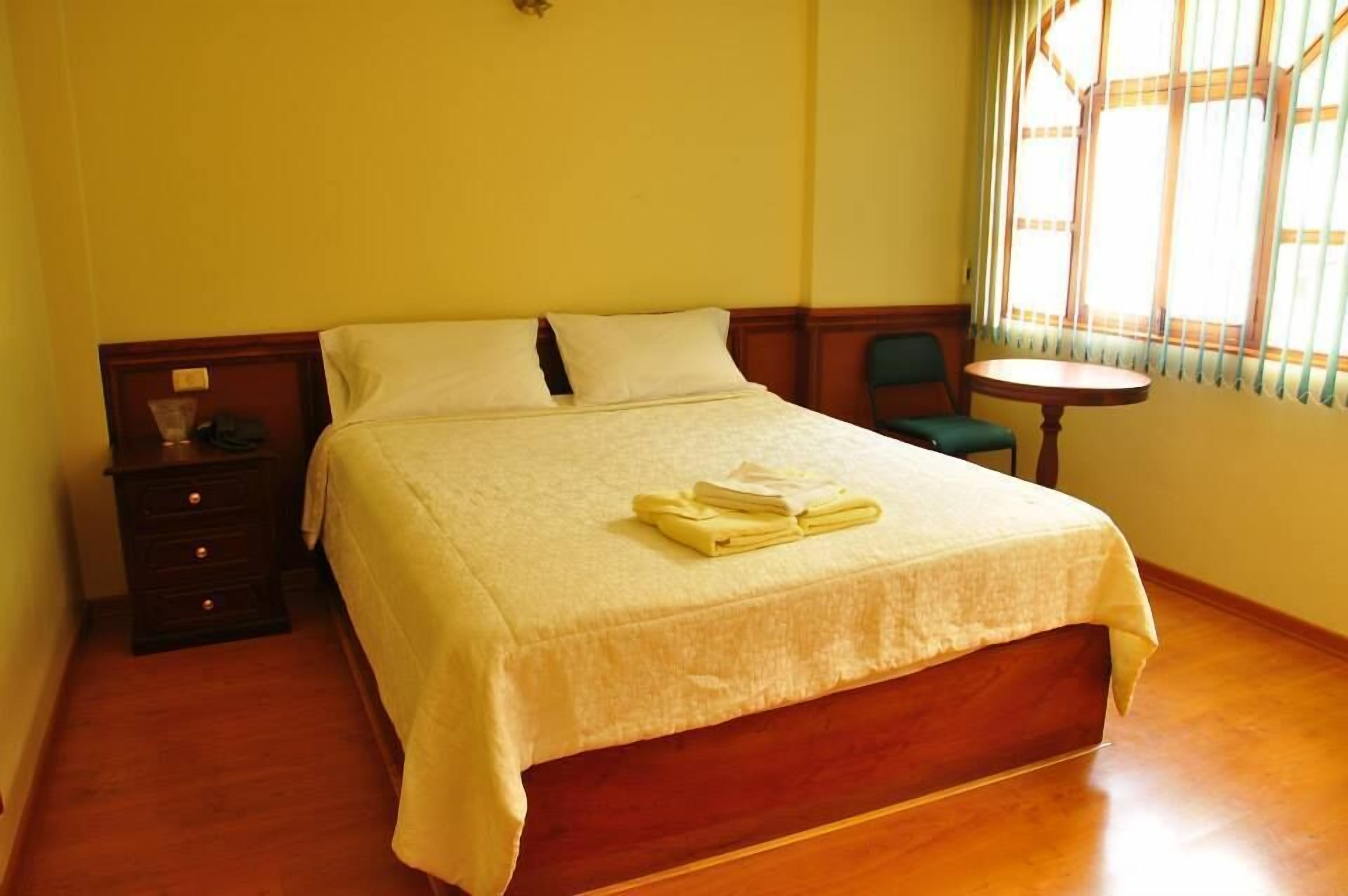 A neatly made bed with a white bedspread, a wooden nightstand with drawers, a small round table, and a window with vertical blinds.
