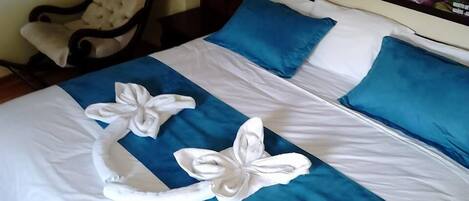 Una cama bien hecha con ropa de cama azul y blanca, dos toallas blancas dispuestas en forma de lazo, una silla y una lámpara montada en la pared.