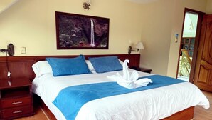 Una cama bien hecha con ropa de cama azul y blanca, cabecera de madera, mesita de noche con lámpara y un cuadro de una cascada en la pared.