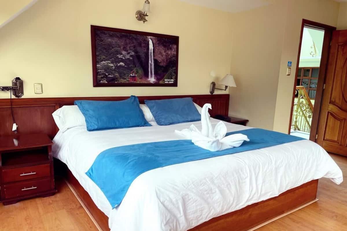 Een netjes opgemaakt bed met blauw-witte beddengoed, een houten hoofdbord, een nachtkastje met een lamp, en een schilderij van een waterval aan de muur.