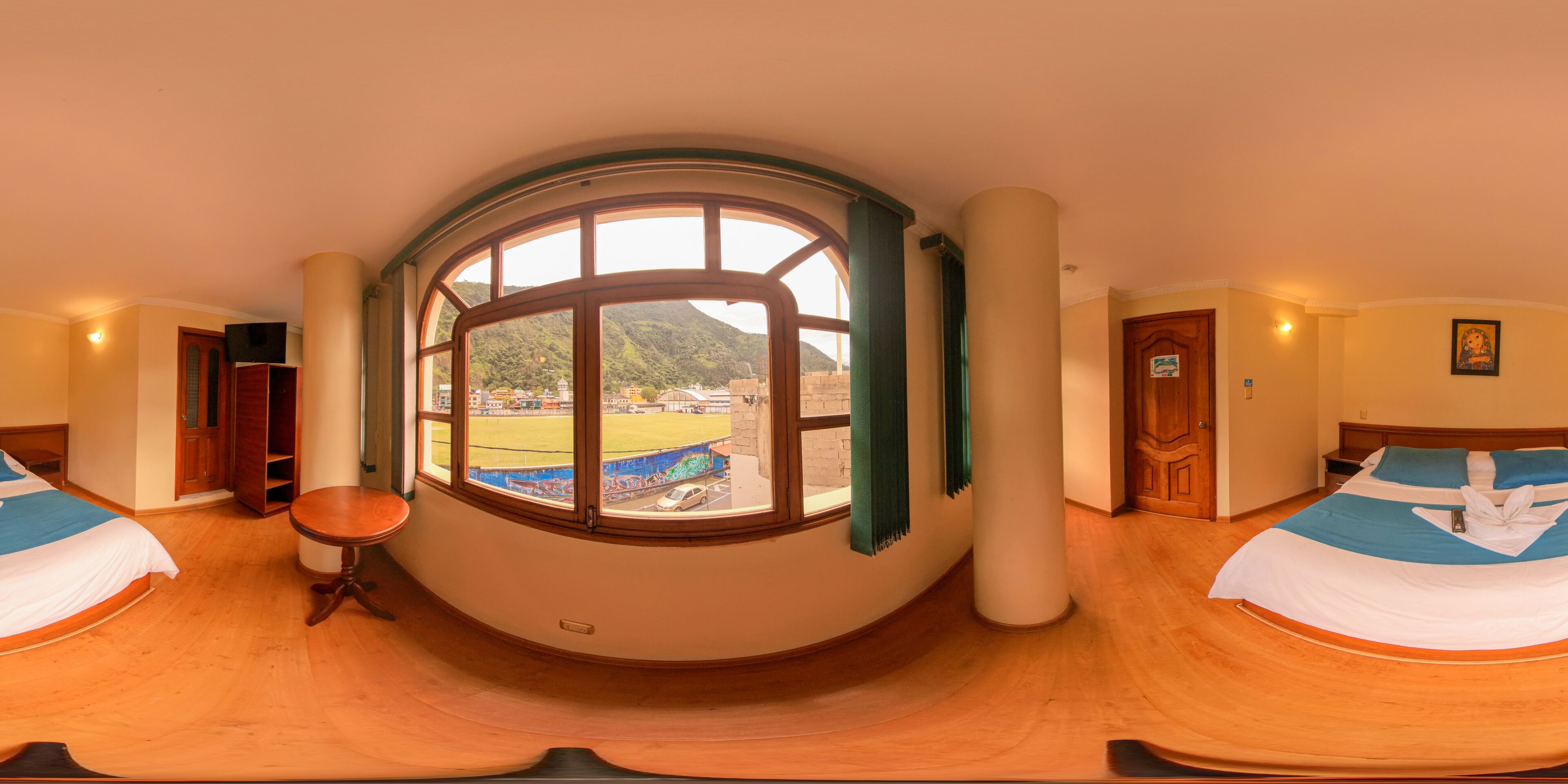 A room with a large arched window, two beds with blue and white bedding, a round wooden table, and a view of a mountainous landscape.