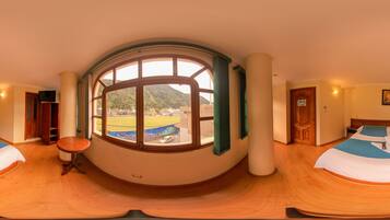 A room with a large arched window, two beds with blue and white bedding, a round wooden table, and a view of a mountainous landscape.