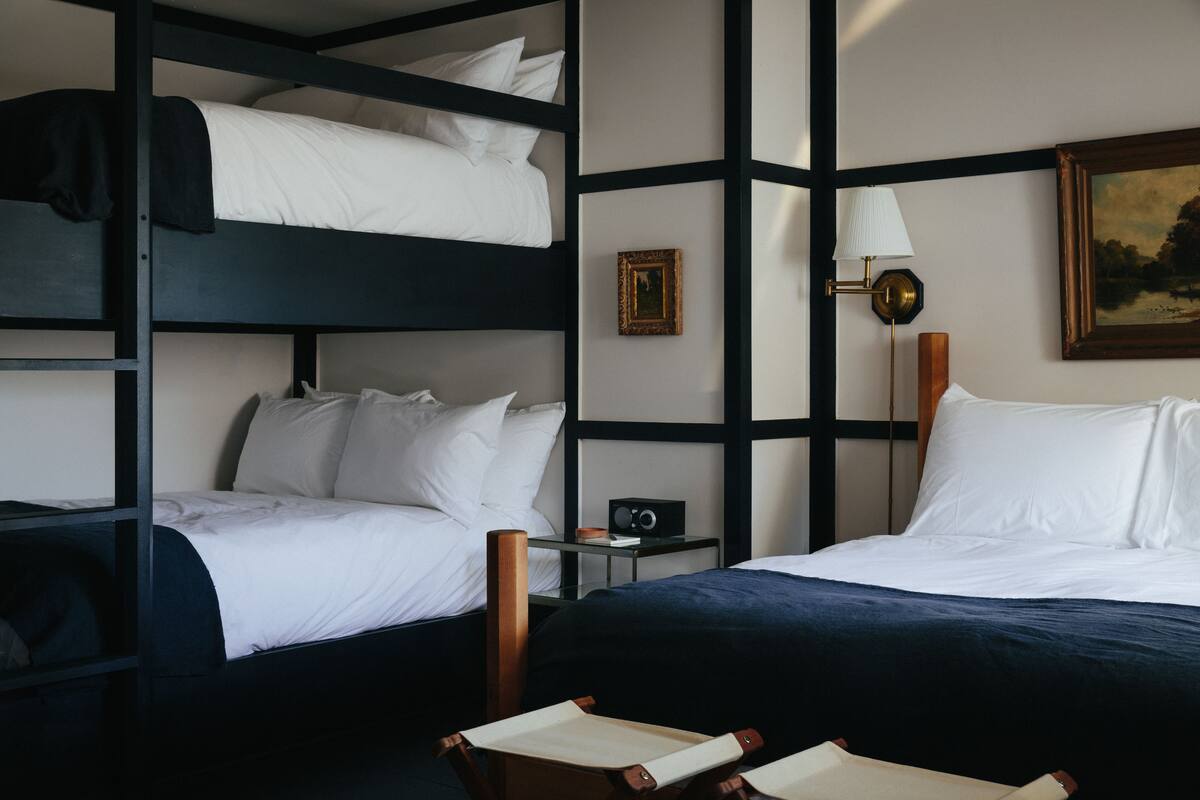 A room with bunk beds, a bedside table with a lamp, a framed picture on the wall, and a chair.